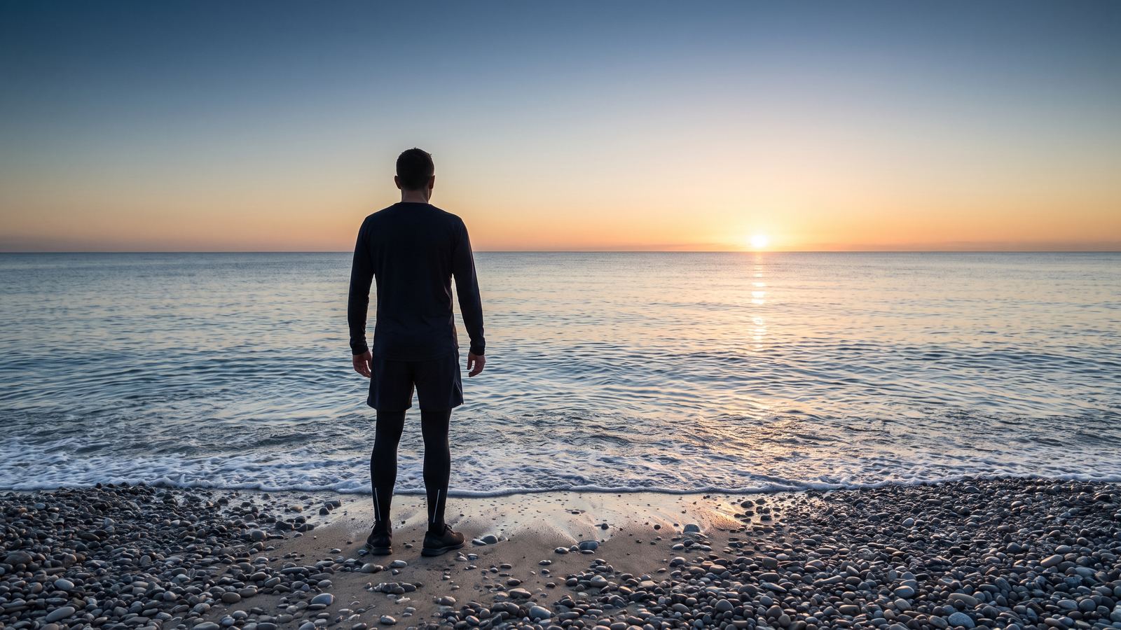An athlete in dark training clothes stands at the water's edge on a pebble beach at sunrise, looking out over a calm sea towards the sun on the horizon